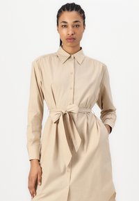 Vestito camicia beige con fronte a bottoni, colletto piegato e maniche lunghe. Dettaglio vita legata e cuciture delle spalle gatherate. Tessuto morbido e liscio.