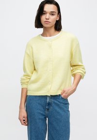 Hele kollane pehme materjaliga kardigan. Omadusteks on ümmargune kaelus, ees nupud ja rullitud varrukasäär. Sobib kokku siniste teksapükstega.