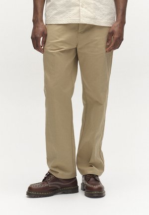 Bărbat purtând pantaloni bej cu picior drept și pantofi maro închis din piele cu șireturi, stând pe un fundal simplu.