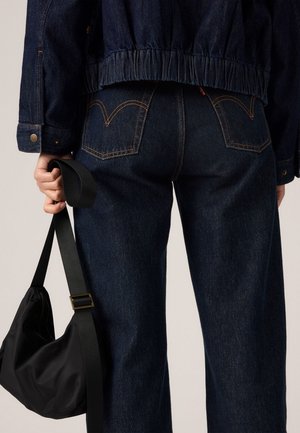 Person med mørkeblå denim jeans og jakke, som holder en sort taske i remmen ved sin side.
