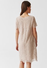 Krajkov dress v jemně béžové barvě s krátkými rukávy, zdobené složitými vzory paisley a vlnitým lemem, ukazující lehký a strukturovaný design.