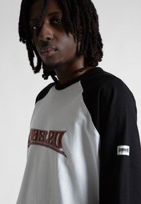 Giovane uomo con dreadlocks che indossa una maglietta raglan bianca e nera con testo grafico rosso sul petto e una toppa sulla manica, sfondo neutro.