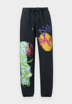 Joven con sudadera negra con capucha y pantalones con diseños de graffiti coloridos, de pie con las manos en los bolsillos contra un fondo blanco liso.