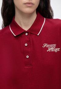 Personne portant un polo rouge avec une bordure blanche au col et un texte brodé blanc "Tommy Hilfiger" sur la poitrine.
