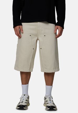 Mężczyzna ubrany w beżowe szorty do kolan, białe skarpety do kostki, czarno-białe sneakersy oraz czarną bluzę z długim rękawem, stojący na jednolitym tle.