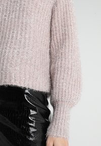 Jasnoróżowy sweter z dzianiny o miękkiej, puszystej teksturze, z prążkowanym wzorem i objętościowymi rękawami, zestawiony z błyszcząca spódnicą z winylu w kolorze czarnym.