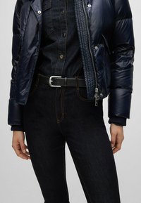Chaqueta acolchada de color marino con textura brillante y acolchada sobre un conjunto de denim oscuro. Cinturón negro con hebilla plateada y costuras naranjas en contraste en los jeans.