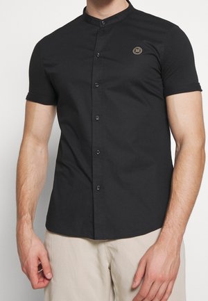 Mężczyzna ubrany w czarną, krótko-rękawkową koszulę zapinaną na guziki z kołnierzem typu mandarynka oraz beżowe spodnie, stojący na jasnym, jednolitym tle.