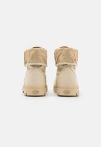 Botas de tobillo de lona beige con un puño plegable, suela de goma y un parche de logo redondo. Presentan broches de metal y un material superior texturizado.