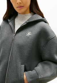 Haut à capuche zippé gris foncé en tissu doux, avec une poche kangourou et un petit logo brodé sur la poitrine. Poignets côtelés.