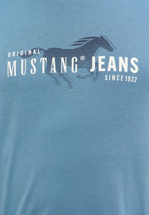 Niebieska koszulka z napisem "ORIGINAL MUSTANG JEANS" na przodzie w odważnym białym kolorze, z stylizowaną grafiką konia powyżej. Miękka, gładka tekstura materiału.