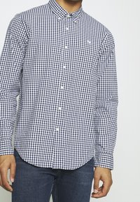 Granatowo-biały koszula w kratkę z guzikiem, z kołnierzykiem typu spread, długimi rękawami i zaokrąglonym wykończeniem. Posiada białe guziki i subtelny detal z brandingiem.