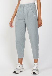 Vaaleanharmaat vakosamettihousut, joissa on rento malli, etupleaatit, taskut ja pieni logo-patch sivussa, yhdistettynä valkoisiin lenkkiareihin.