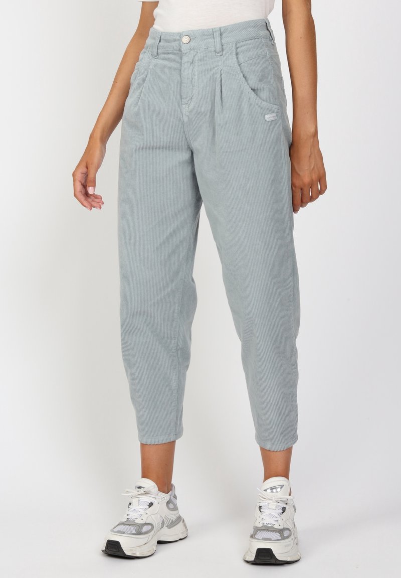 Vaaleanharmaat vakosamettihousut, joissa on rento malli, etupleaatit, taskut ja pieni logo-patch sivussa, yhdistettynä valkoisiin lenkkiareihin.