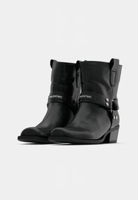 Botine negre din piele, cu toc mic și vârf rotunjit. Au o curea laterală cu butoane argintii și un logo, cu o suprafață texturată.