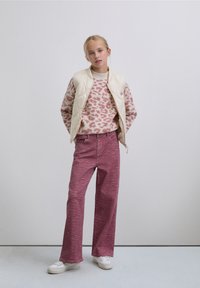 Gesteppte elfenbeinfarbene Weste, rosa Pullover mit Leopardenmuster und weit geschnittene Hose mit roten Streifen, kombiniert mit weißen Turnschuhen.