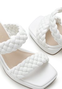 Witte sandalen hebben gevlochten banden en een chunky vierkante hak. De zool is glad, en het interieur heeft een neutraal gekleurd voering.