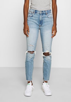 Person wearing light blue ripped jeans and white sneakers, standing against a plain white background.