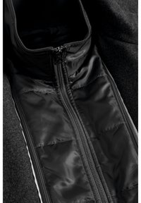 Gros plan d’une section de veste matelassée noire avec une fermeture éclair centrale et un col montant sur un fond en tissu foncé.
