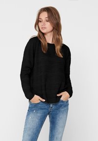 Pull noir avec un tissu texturé, décolleté large et manches longues, associé à un jean bleu clair déchiré. Design simple et décontracté.