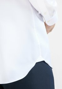 Camicia bianca in tessuto intrecciato con maniche lunghe, caratterizzata da un orlo curvo. Abbinata a pantaloni blu scuro, con attenzione alla texture e alla struttura del tessuto.