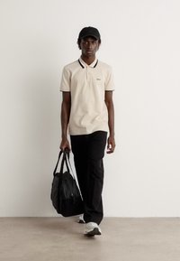 Beżowa koszulka polo z czarnymi akcentami, czarne spodnie i czarna czapka. Osoba niesie ciemną torbę i ma na sobie białe trampki.