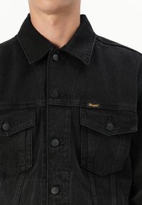 Primer plano de una persona que lleva una chaqueta Wrangler de mezclilla negra con bolsillos en el pecho con botones y botones metálicos con la marca.