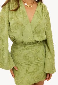 Robe verte texturée avec un décolleté en V et des manches longues. Le tissu présente un motif à franges et une silhouette ajustée. Des bijoux en or accentuent l'ensemble.
