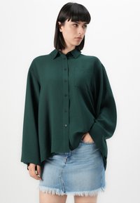 Mørk grønn oversized skjorte med krage og frontlomme, sammen med en lys blå denim miniskjørt med frayed kant.