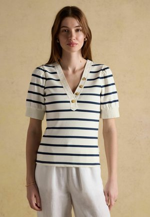 Frau trägt ein weißes Kurzarmoberteil mit schwarzen horizontalen Streifen und goldenen Knöpfen, kombiniert mit lockeren weißen Hosen vor einem beigen Hintergrund.