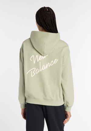 Persoon met gevlochten haar, gekleed in een lichtgroene hoodie met de tekst "New Balance" op de achterkant en zwarte broek, met de rug naar de kijker gericht.