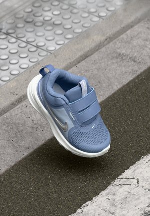 Chaussure de sport bleue avec tige en maille, deux sangles Velcro, accents argentés et semelle blanche. Conçue pour le confort et la stabilité.