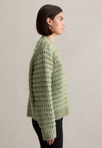 Grüner Strickpullover mit texturiertem, offenem Maschenmuster, geripptem Saum und Bündchen. Cropped-Länge, getragen mit schwarzen Hosen.