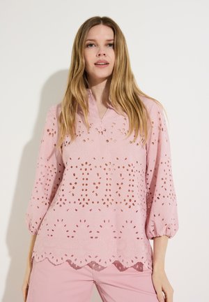 Frau mit langen blonden Haaren, die eine hellrosa Lochbluse und passende rosa Hose trägt, stehend vor einem schlichten weißen Hintergrund.