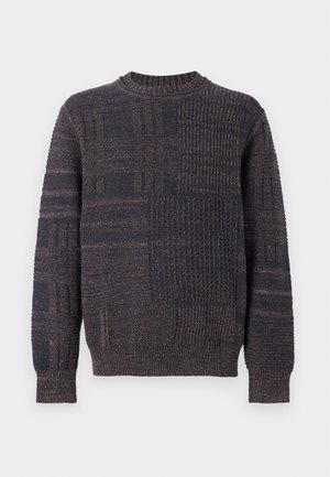 Homme portant un pull en maille texturée foncée, un pantalon large marron et des baskets marron, debout devant un fond clair uni.