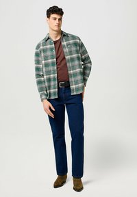 Camisa a cuadros verde y blanca sobre una camiseta marrón, combinada con unos vaqueros azul oscuro y botas marrones. La camisa cuenta con cierres de botones y mangas largas.