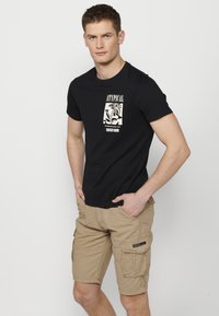 Melna kokvilnas T-kreklu ar grafisku druku, kombinēts ar bēšiem kravas šortiem ar vairākām kabatām un mazu zīmola etiketīti sānos.