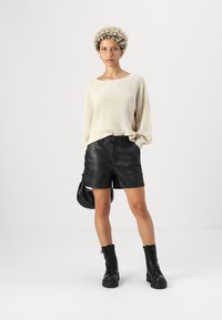 Helle beige, übergroße Strickjacke mit weitem Halsausschnitt, kombiniert mit schwarzen Leder-Shorts. Das Model trägt schwarze Kampfboots und hält eine kleine Tasche.
