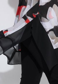 Fließender, transparenter schwarzer Stoff mit Muster aus roten, weißen und grauen abstrakten Formen über enganliegenden schwarzen Hosen.