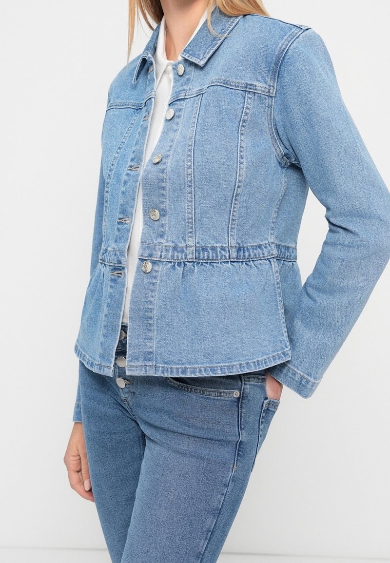 Femme portant une veste en jean bleu clair avec des boutons et un jean assorti, debout avec une main dans la poche devant un fond uni.
