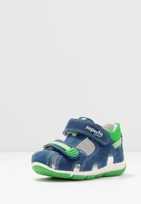 Sininen mokkasiinisandaali vihreillä yksityiskohdilla, säädettävä hihna, avoimet sivut ilmanvaihtoa varten ja teksturoitu kumipohja. Hihnassa on logo.