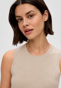 Beige mouwloze gebreide top met geribbelde halslijn en armsgaten, met een gladde textuur en een getailleerde vorm, gedragen door iemand met kort haar.