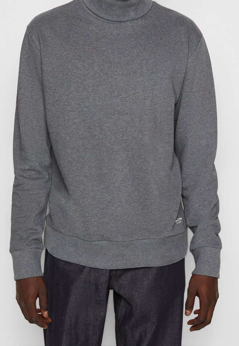 Homme portant un pull à col roulé gris uni à manches longues et un jean denim foncé, les mains détendues le long du corps.