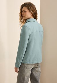 Giacca di cotone azzurro chiaro con colletto, maniche lunghe e polsini con bottoni. Ha un taglio dritto e una texture sottile. Indossata con pantaloni grigi sbiaditi.