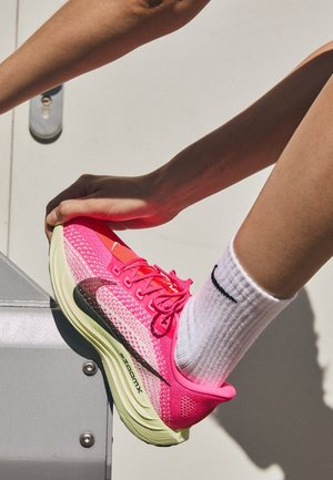 Persoon met witte Nike-sok en felroze Nike ZoomX hardloopschoen die het been strekt op een metalen oppervlak.
