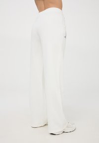 Osoba w luźnych, białych spodniach z gwiazdkowym wzorem po boku, w komplecie z białymi butami sportowymi, stoi na jednolitym tle.