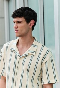 Short-sleeve shirt in cream with vertical green stripes, featuring a collar, button closure, and textured fabric.