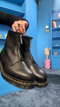 Άτομο κρατάει ένα ζευγάρι μαύρα δερμάτινα μποτάκια με φερμουάρ και κίτρινα ράμματα, σε ένα μπλε δωμάτιο με ράφια και διακοσμητικά αντικείμενα.