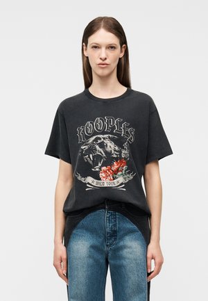 Γυναίκα με μαύρο μπλουζάκι με σχέδιο μοβ επικεφαλής πάνθηρα και κόκκινα λουλούδια, συνδυασμένο με δίχρωμο μπλε τζιν, στέκεται μπροστά σε απλό φόντο.