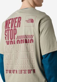 Béžové bavlněné tričko s výrazným vínovým textem "NEVER STOP EXPLORING" a souřadnicemi tištěnými na zadní straně; obsahuje malé logo v horním rohu.